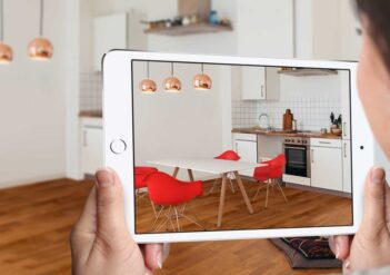 fotografia com uma mulher segurando um tablet, que mostra uma sala com paredes brancas, piso de madeira e cadeiras vermelhas