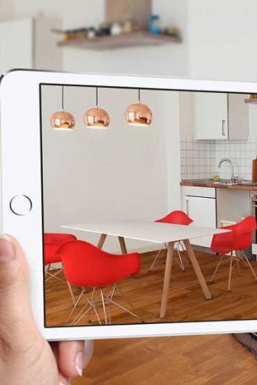 fotografia com uma mulher segurando um tablet, que mostra uma sala com paredes brancas, piso de madeira e cadeiras vermelhas