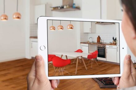fotografia com uma mulher segurando um tablet, que mostra uma sala com paredes brancas, piso de madeira e cadeiras vermelhas