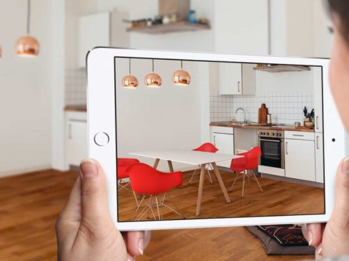 fotografia com uma mulher segurando um tablet, que mostra uma sala com paredes brancas, piso de madeira e cadeiras vermelhas