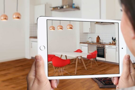 fotografia com uma mulher segurando um tablet, que mostra uma sala com paredes brancas, piso de madeira e cadeiras vermelhas