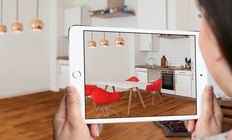fotografia com uma mulher segurando um tablet, que mostra uma sala com paredes brancas, piso de madeira e cadeiras vermelhas