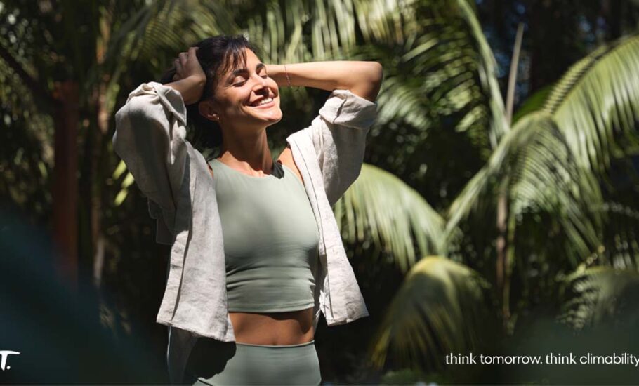 Fotografia de uma mulher sorrindo, com as mãos na cabeça, em frente a palmeiras.