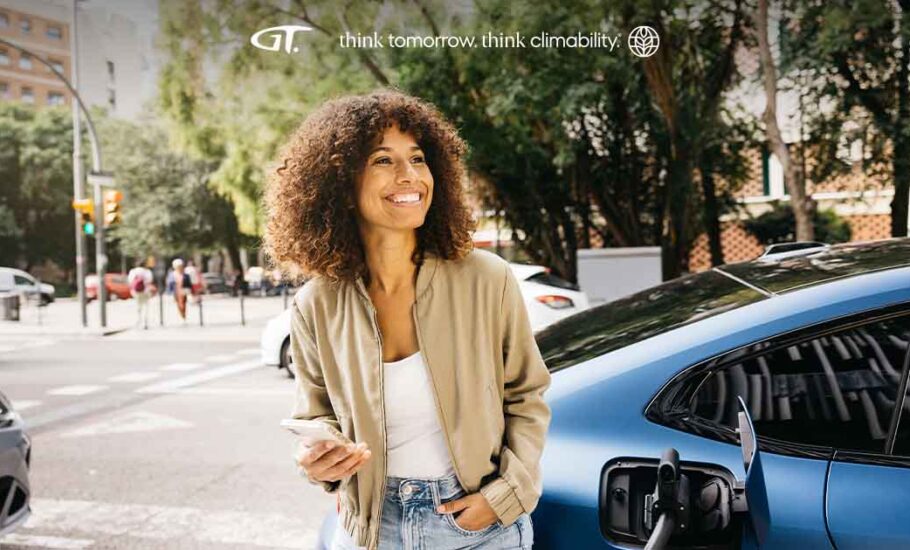Fotografia de uma mulher com pele negra, cabelo cacheado e roupas claras, na rua em frente a um carro
